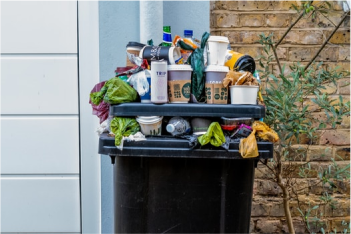 An overflowing black rubbish bin with various packaging placed on top including plastic bags single-use and coffee cups