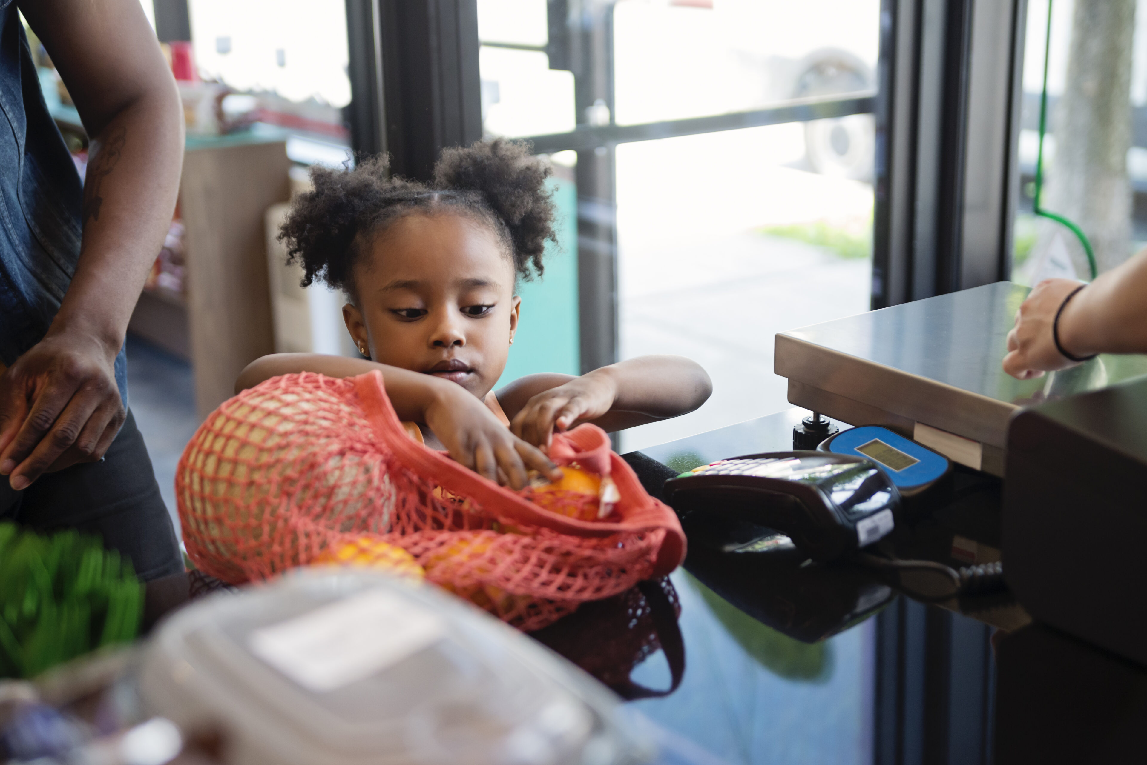 A little girl is stood next to an adult that is out of shot. She is packing their plastic-free shopping into a reusable coral mesh bag