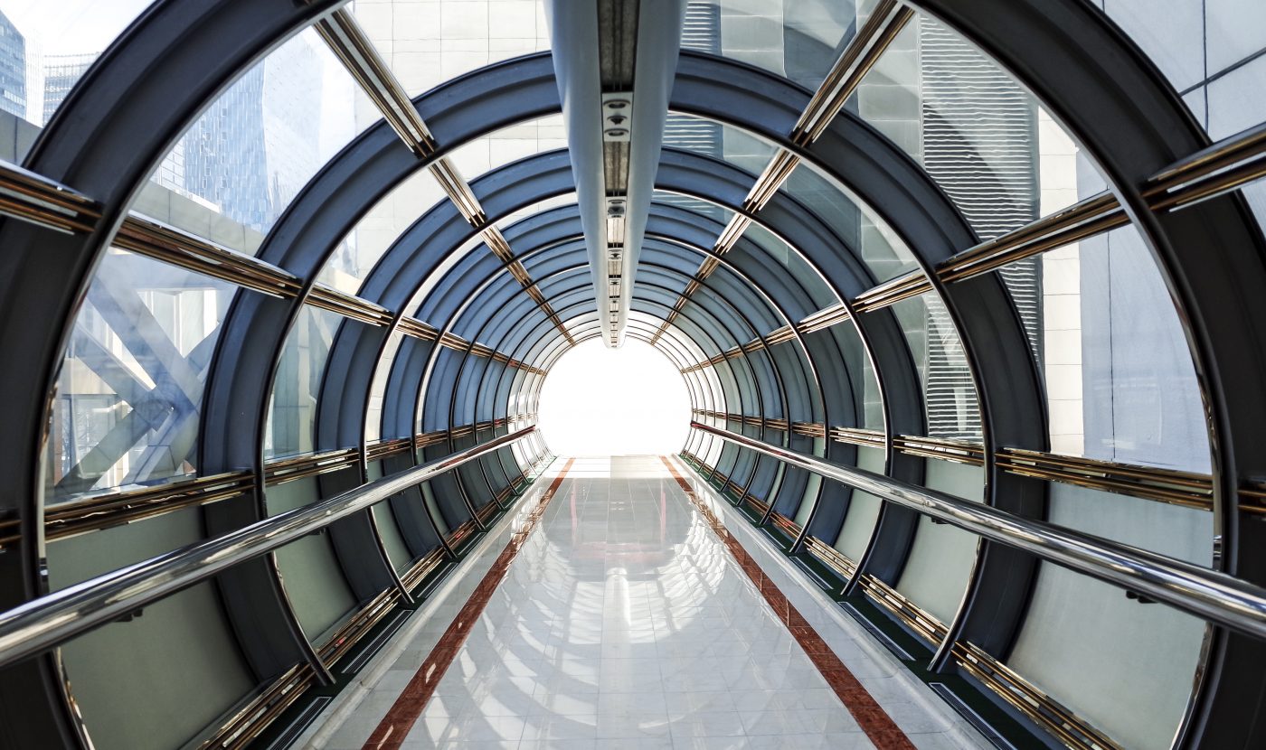 Photograph of a tunnel made from glass and metal leading to a bright light. Tunnel is surrounded by commercial buildings in the city of London